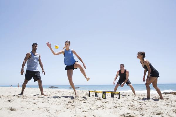 Vier Personen spielen Spikeball am Strand bei klarem Himmel und Meer im Hintergrund.