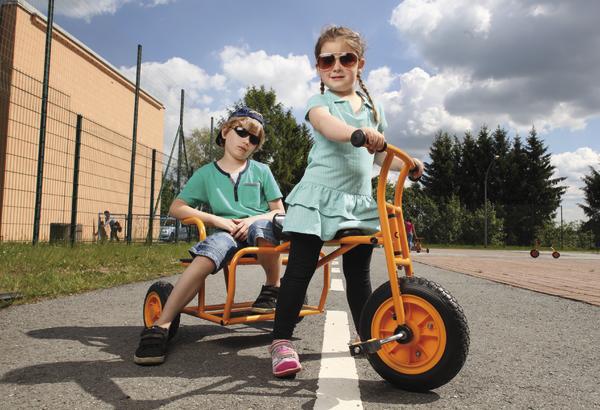 Kinder fahren auf einem großen orangenen Dreirad auf einem sonnigen Spielplatz.