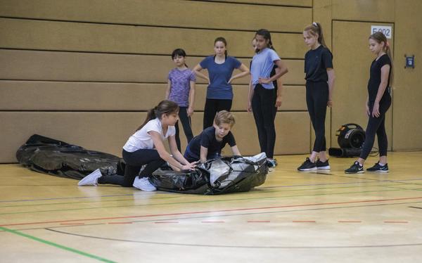 Kinder in Sportkleidung rollen eine Matte in einer Turnhalle, andere beobachten.