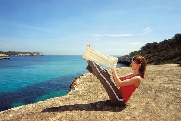 Frau liest Buch am Felsen mit Blick auf das blaue Meer und klaren Himmel.