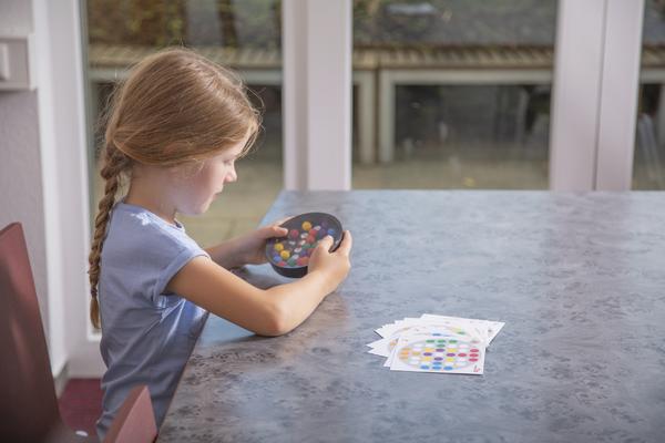 Mädchen spielt mit bunten Spielsteinen und Karten an einem Tisch im hellen Raum.