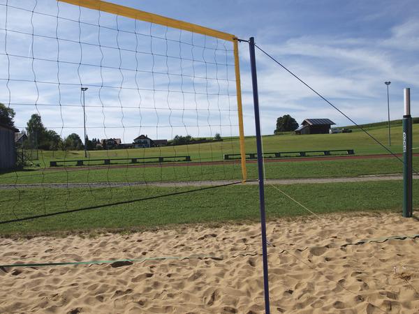 Beachvolleyballnetz auf Sandplatz mit grüner Wiese und blauem Himmel im Hintergrund.
