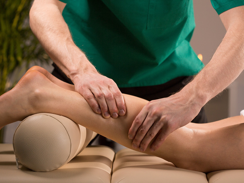Patient mit untergelegter Lagerungsrolle wird vom Therapeut an der Wade behandelt.