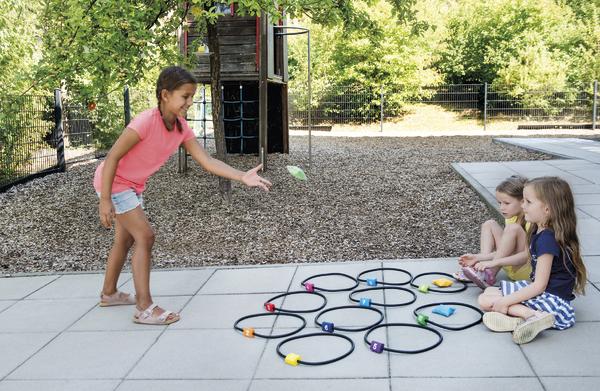 Kind wirft Beanbag auf Zielringe, zwei Kinder beobachten, draußen auf Spielplatz im Sommer