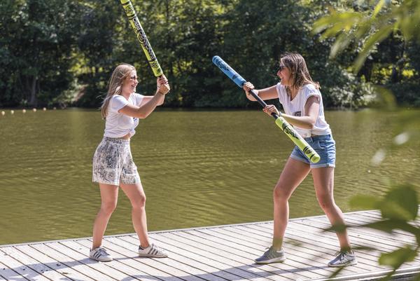 Zwei junge Frauen spielen mit Schaumstoffschlägern auf einem Steg am See im Sommer.