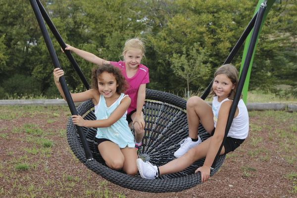 Drei Mädchen spielen lachend auf einer großen Nestschaukel im Park bei Tageslicht.