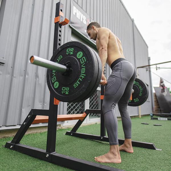 Mann trainiert barfuß mit Langhantel im Freien vor Fitnessstudio.