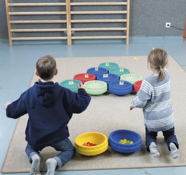 Zwei Kinder knien in einer Turnhalle und werfen kleine Bälle auf nummerierte Schaumstoffscheiben auf einer Matte.