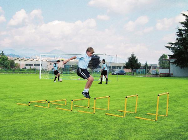 Junge trainiert Sprungkraft mit Hürden auf Fußballplatz, andere Spieler im Hintergrund.