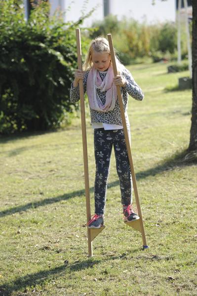 Mädchen balanciert auf Holzstelzen im Garten bei sonnigem Wetter.