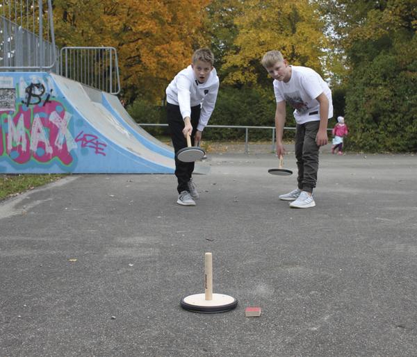 Zwei Jungen spielen Wikingerschach auf einem Skatepark mit herbstlichen Bäumen im Hintergrund.