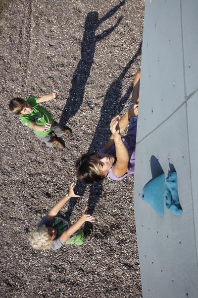 Kinder klettern an einer Kletterwand mit Sicherung durch Freunde auf Kiesboden.