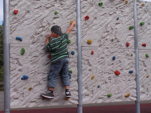 Kind klettert an bunter Kletterwand im Freien, trägt gestreiftes Shirt und Cap.