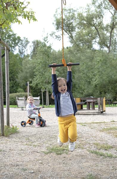 Kind schwingt an Seil im Park, anderes Kind fährt Dreirad im Hintergrund, sonniger Tag.