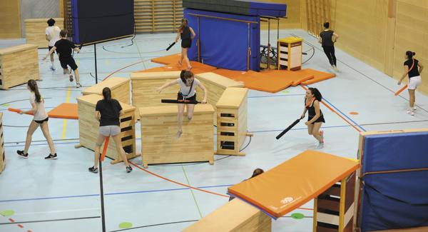 Gruppe von Personen spielt in einer Turnhalle ein Parcours-Spiel mit Schaumstoffstäben und Hindernissen.
