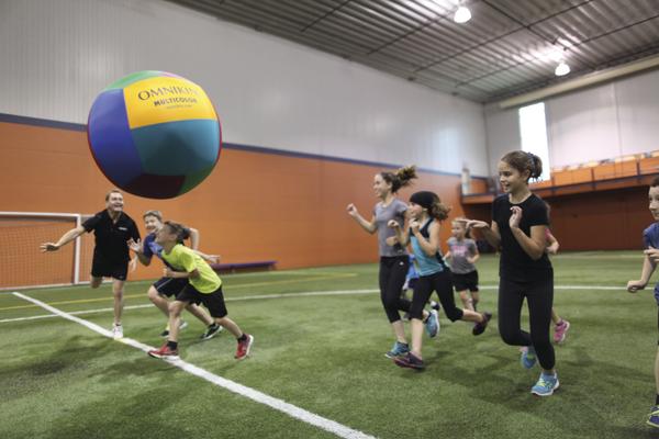 Kinder spielen mit einem großen bunten Ball in einer Indoor-Sporthalle auf Kunstrasen.