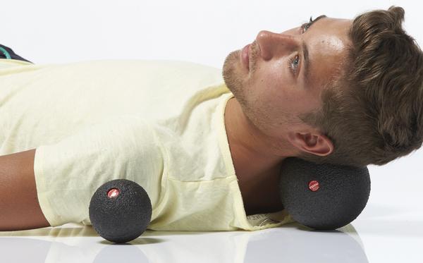 Mann liegt auf dem Rücken und verwendet einen Massageball im Nackenbereich zur Entspannung der Muskulatur, während ein weiterer Ball daneben liegt.