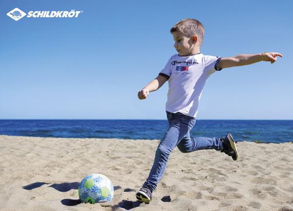 Kleiner Junge schießt einen Ball am Strand, im Hintergrund ist das Meer zu sehen, das Schildkröt-Markenlogo ist sichtbar.