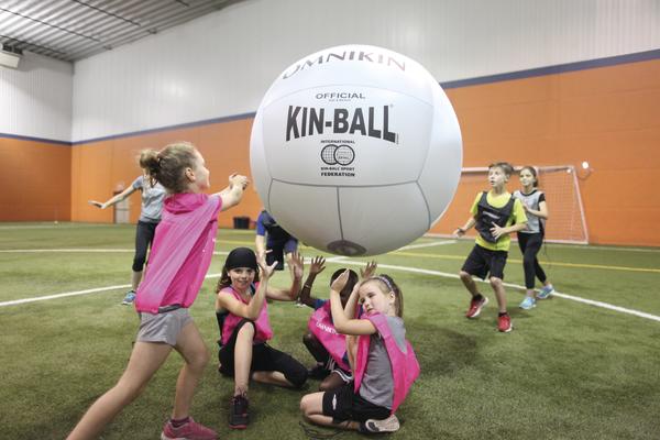 Kinder spielen Kin-Ball in einer Halle auf Kunstrasen mit großem weißem Ball.
