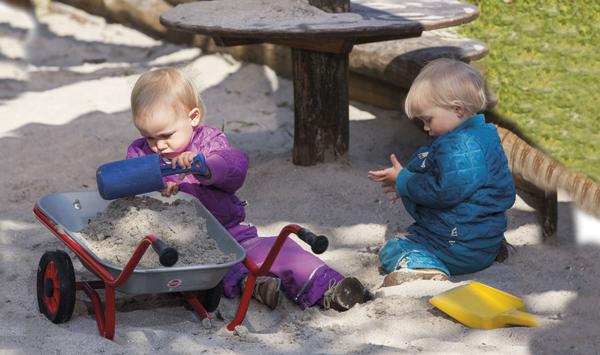 Zwei Kleinkinder spielen im Sandkasten mit Schaufel und Schubkarre bei sonnigem Wetter.