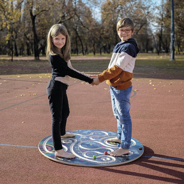 Zwei Kinder halten sich an den Händen und stehen barfuß auf einem bunten Spielbrett im Freien.
