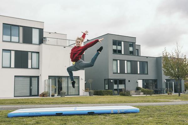 Frau springt auf Luftmatratze im Garten vor modernen Häusern bei bewölktem Himmel.