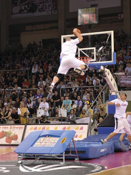 Basketballspieler springt mit Trampolin zum Dunking vor vollem Publikum in Halle.