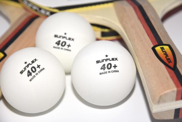 Drei weiße Sunflex Tischtennisbälle und zwei Holztischtennisschläger auf weißem Hintergrund.