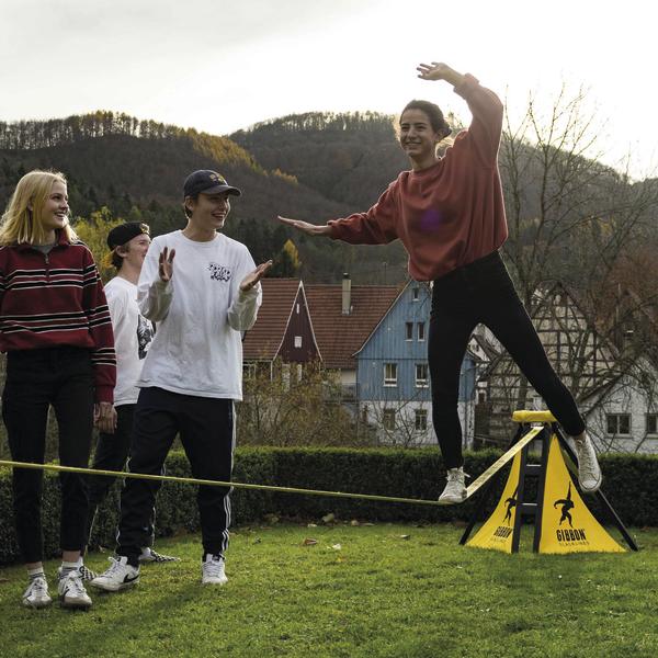 Jugendliche balancieren auf Slackline im Garten vor Häusern und Hügeln bei Sonnenuntergang.