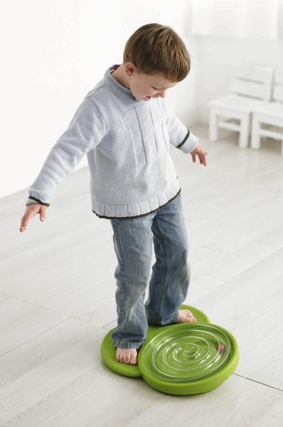 Junge balanciert barfuß auf grünem Balance-Board in hellem Raum mit Holzboden.