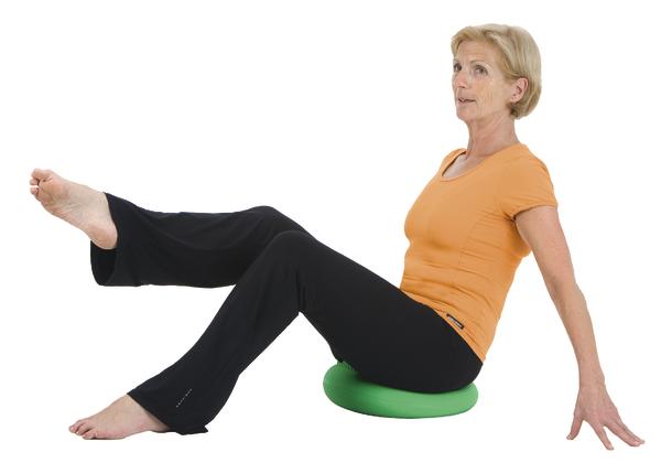 Frau macht Gleichgewichtsübung auf grünem Balancekissen, trägt orange Shirt und schwarze Hose.