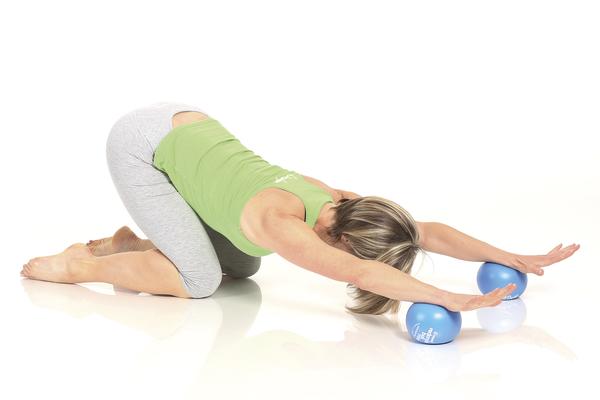 Frau macht Dehnübung mit Pilates-Bällen auf weißem Boden, grüne Sportkleidung.