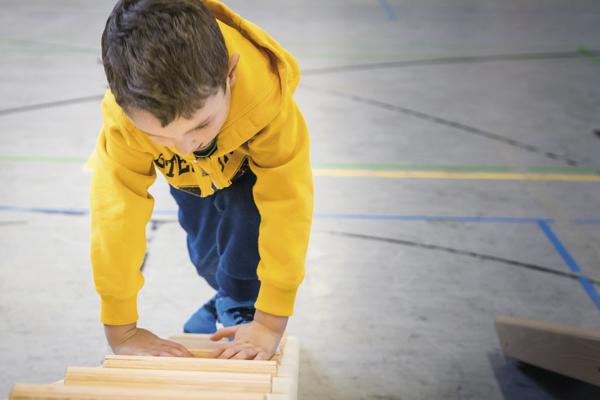 Junge in gelbem Hoodie klettert auf Holzklettergerüst in Turnhalle.