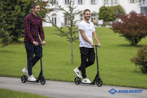 Zwei Männer fahren mit E-Scootern auf einem Gehweg im Park bei Sonnenschein.