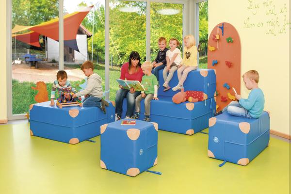 Kinder sitzen auf blauen Sitzwürfeln und hören einer Frau beim Vorlesen in einem hellen Spielzimmer zu.