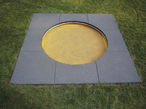 Eingelassene Bodentrampolin mit gelber Sprungfläche und grauen Randplatten im Garten.