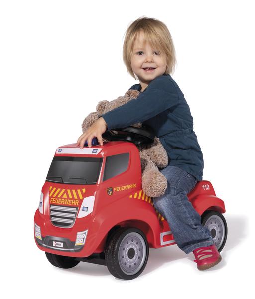 Kind sitzt auf rotem Feuerwehr-Spielzeugauto mit Teddybär, weißer Hintergrund.