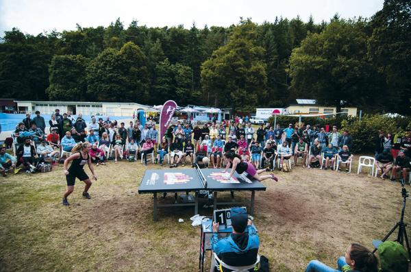 Outdoor-Tischtennisturnier mit Zuschauern vor Waldkulisse bei sonnigem Wetter.