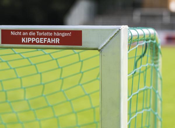 Warnschild "Nicht an die Torlatte hängen! Kippgefahr" an einem Fußballtor mit grünem Netz.