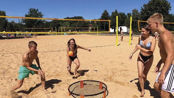 Jugendliche spielen Spikeball am sonnigen Sandstrand mit Volleyballnetzen im Hintergrund.