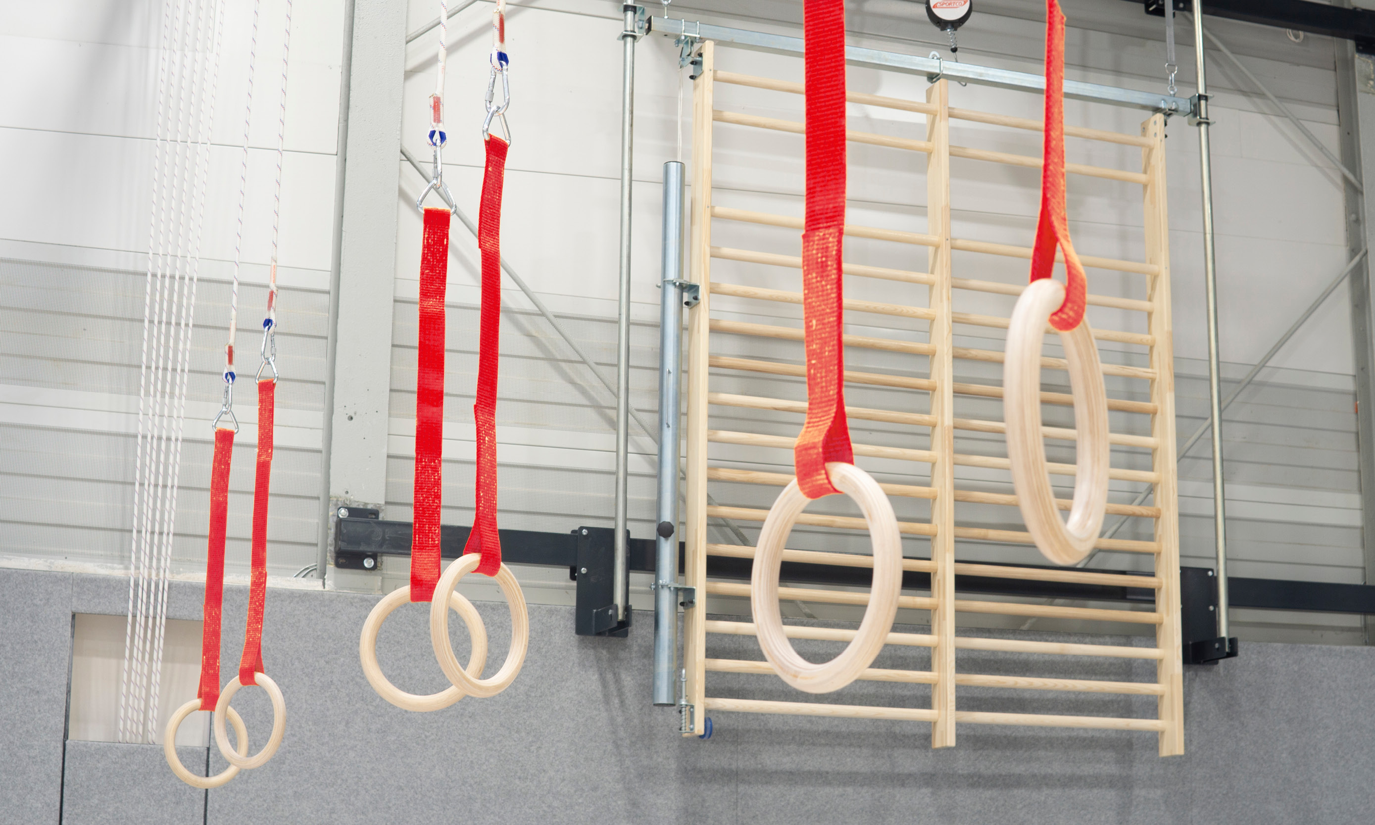 Hängende Holz-Schaukelringe mit roten Sicherheitsseilen in einer Turnhalle, im Hintergrund eine Sprossenwand.