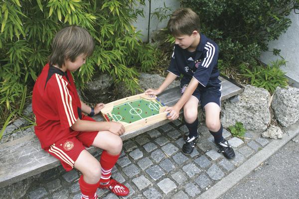 Zwei Jungen in Fußballtrikots spielen Tischfußball draußen auf einer Bank.