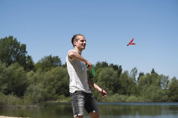 Junger Mann wirft roten Frisbee am Seeufer bei sonnigem Wetter.