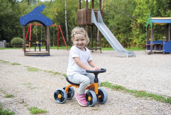 Kleines Mädchen fährt lachend auf orangem Laufrad im Spielplatz mit Rutsche und Schaukel.