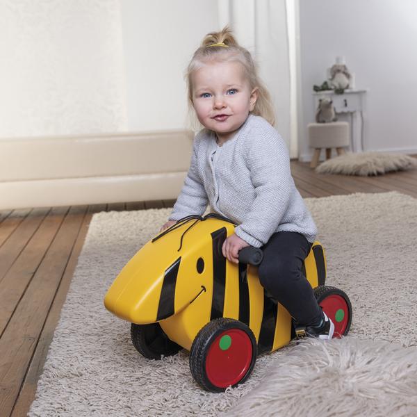 Kleines Mädchen sitzt auf gelbem Kinderfahrzeug in Bienen-Design auf Teppich im Wohnzimmer.