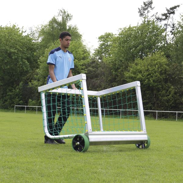 Junger Mann schiebt mobiles Fußballtor auf grünem Sportplatz mit Bäumen im Hintergrund.