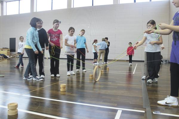 Kinder spielen in der Turnhalle ein Geschicklichkeitsspiel mit Holzringen und Seilen.