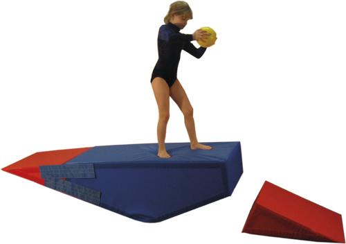 Mädchen in Gymnastikanzug steht auf blau-rotem Schaumstoffspielzeug und hält gelben Ball.