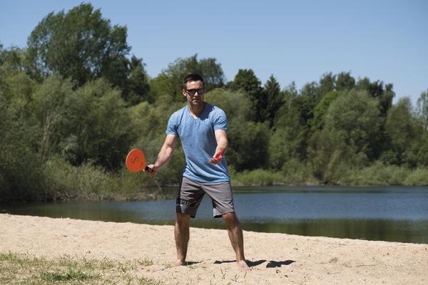 Mann spielt Beach-Tennis am Seeufer bei sonnigem Wetter mit grüner Natur im Hintergrund.