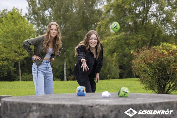 Zwei junge Frauen spielen draußen das Wurfspiel "Schildkröt" auf einer Steintischplatte.